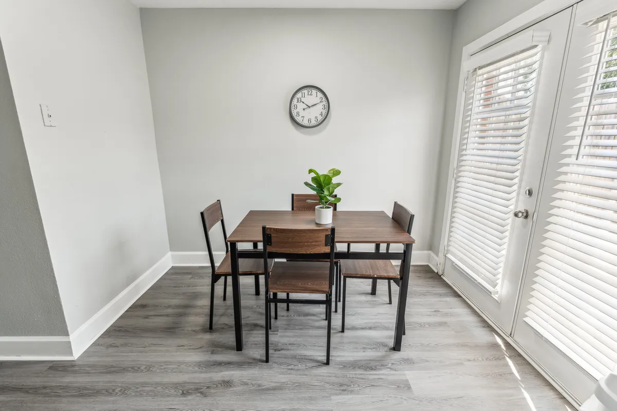 A cozy dining area with a large table surrounded by chairs, a potted plant centerpiece, wall clock, and natural light streaming through a sliding door. The arrangement is welcoming, suitable for family meals, celebrations, or even a casual coffee or work session. The open layout fosters togetherness and comfortable gatherings.