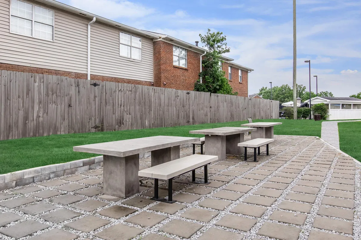 A casual and welcoming communal lawn area with concrete tables and benches, set against a well-maintained green space and enclosed by a fence. The area is designed for outdoor dining, social gatherings, and quiet relaxation, embodying the spirit of both community and solitude.