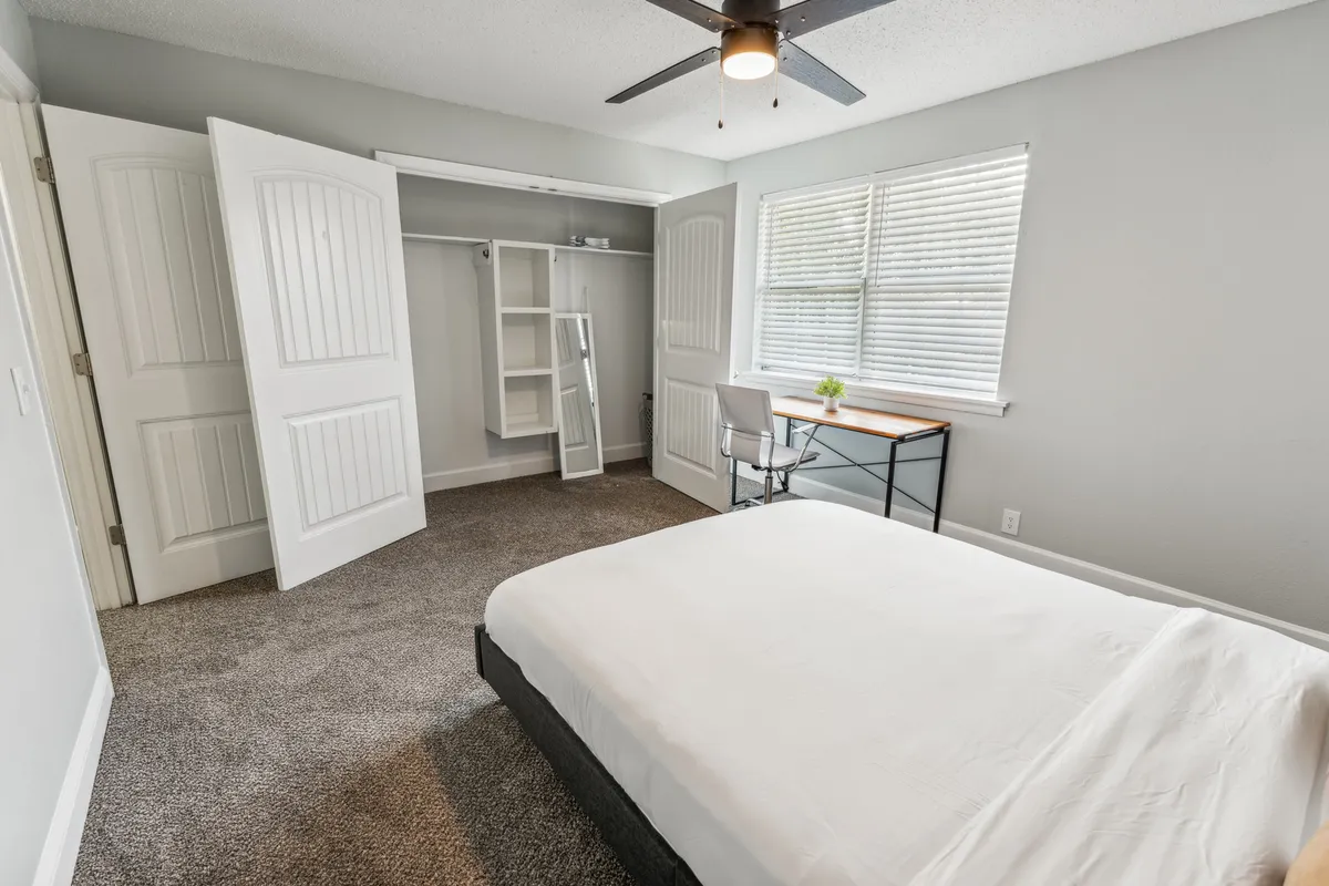 A serene bedroom space featuring a comfortable bed, a desk by the window for working or reading, and a modest closet. Natural light streams in, and the furnishings offer a calming, inviting atmosphere.
