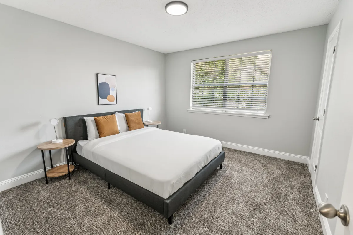 A serene bedroom featuring a bed with soft, decorative pillows, a window letting in gentle natural light, tasteful artwork, and a nightstand with a lamp. The space feels tranquil and designed for rest.