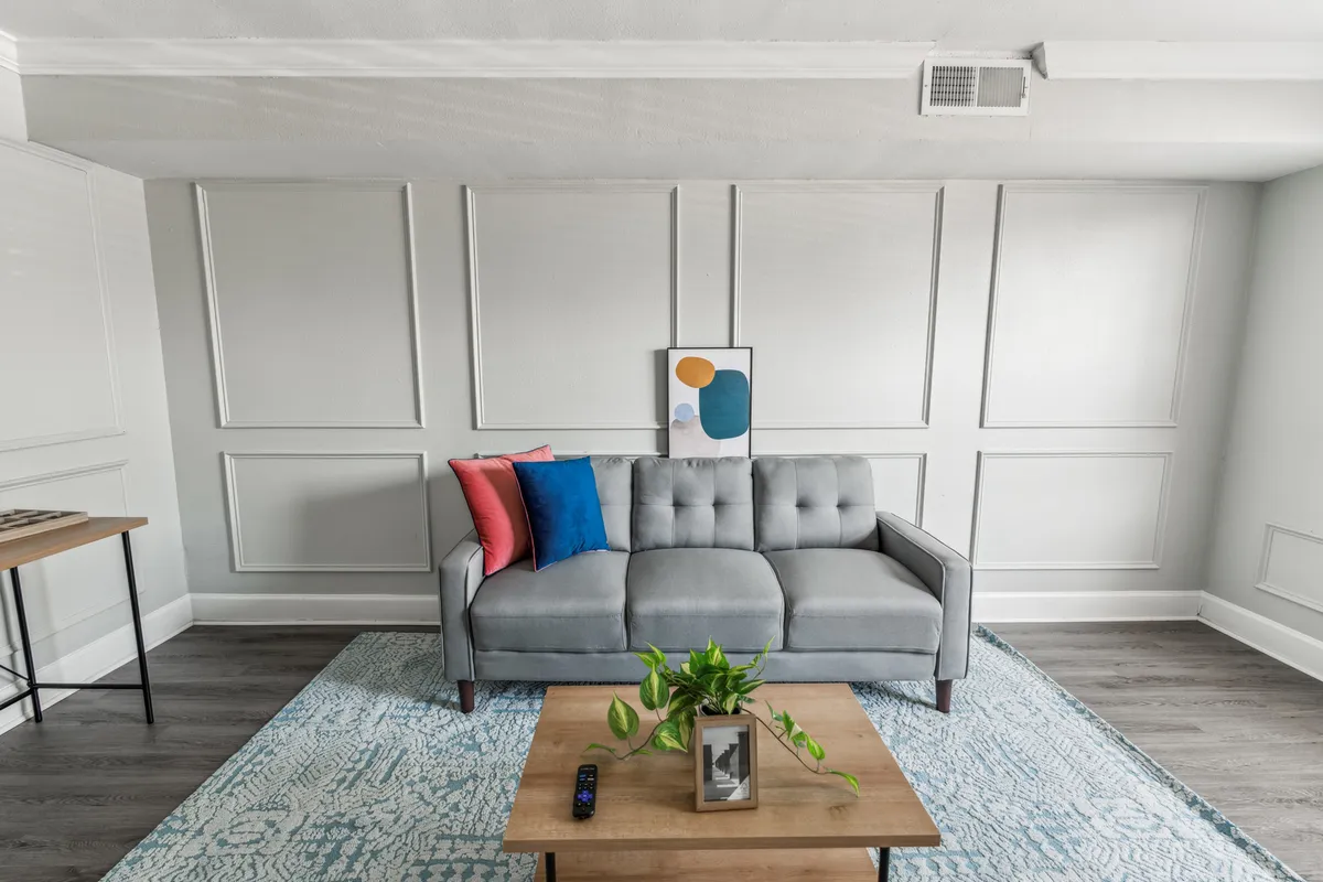 The image showcases a stylish and inviting living room, featuring a plush sofa with chic cushions, a modern coffee table, eye-catching artwork, a soft area rug, and a leafy plant. Ample natural light streams in, accentuating the cozy and calm atmosphere—perfect for relaxing or gathering with friends and family.