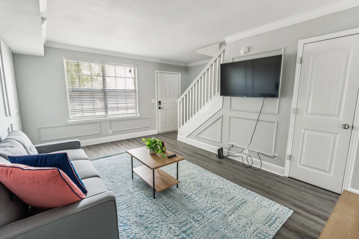 A welcoming living room featuring a comfortable sofa with colorful cushions, a sleek coffee table, a wall-mounted TV, and a staircase in the background. Ample natural light streams in, highlighting contemporary décor and inviting seating.