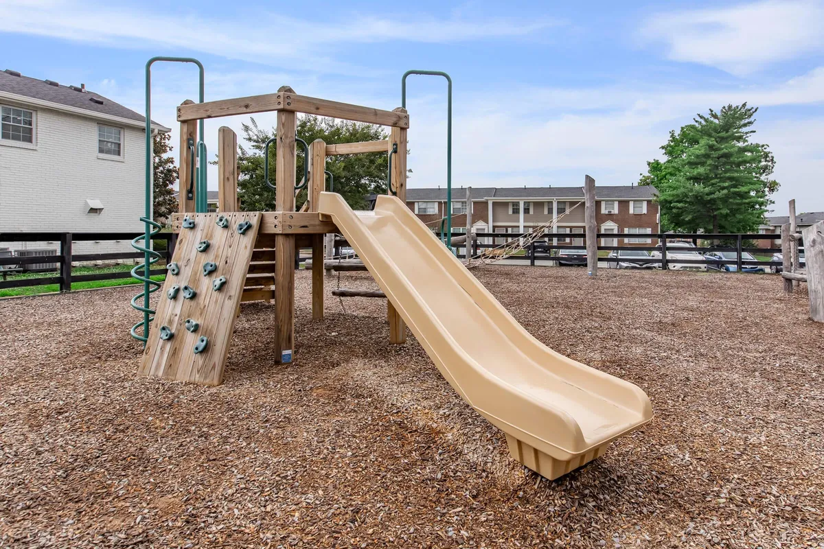This image features vibrant playground equipment, including a slide and climbing wall, with children actively playing and families gathered nearby. The scene is filled with joyful, energetic activity, highlighting a family-friendly neighborhood atmosphere.