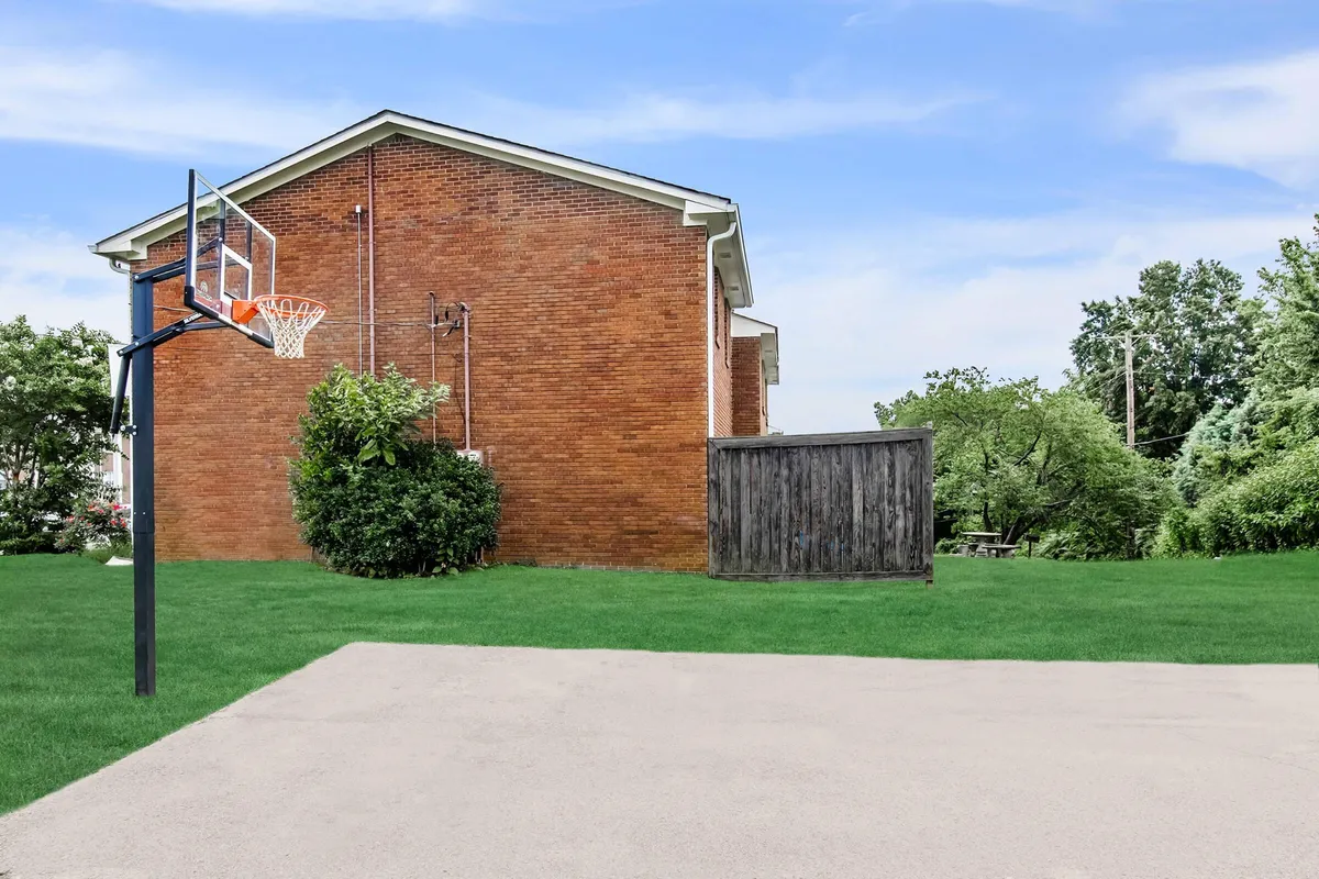 The image displays an outdoor basketball hoop set against a manicured lawn and a brick wall, suggesting a communal recreational area. The open, landscaped space invites outdoor activities and gatherings, giving a sense of connection to neighborhood energy and leisure.